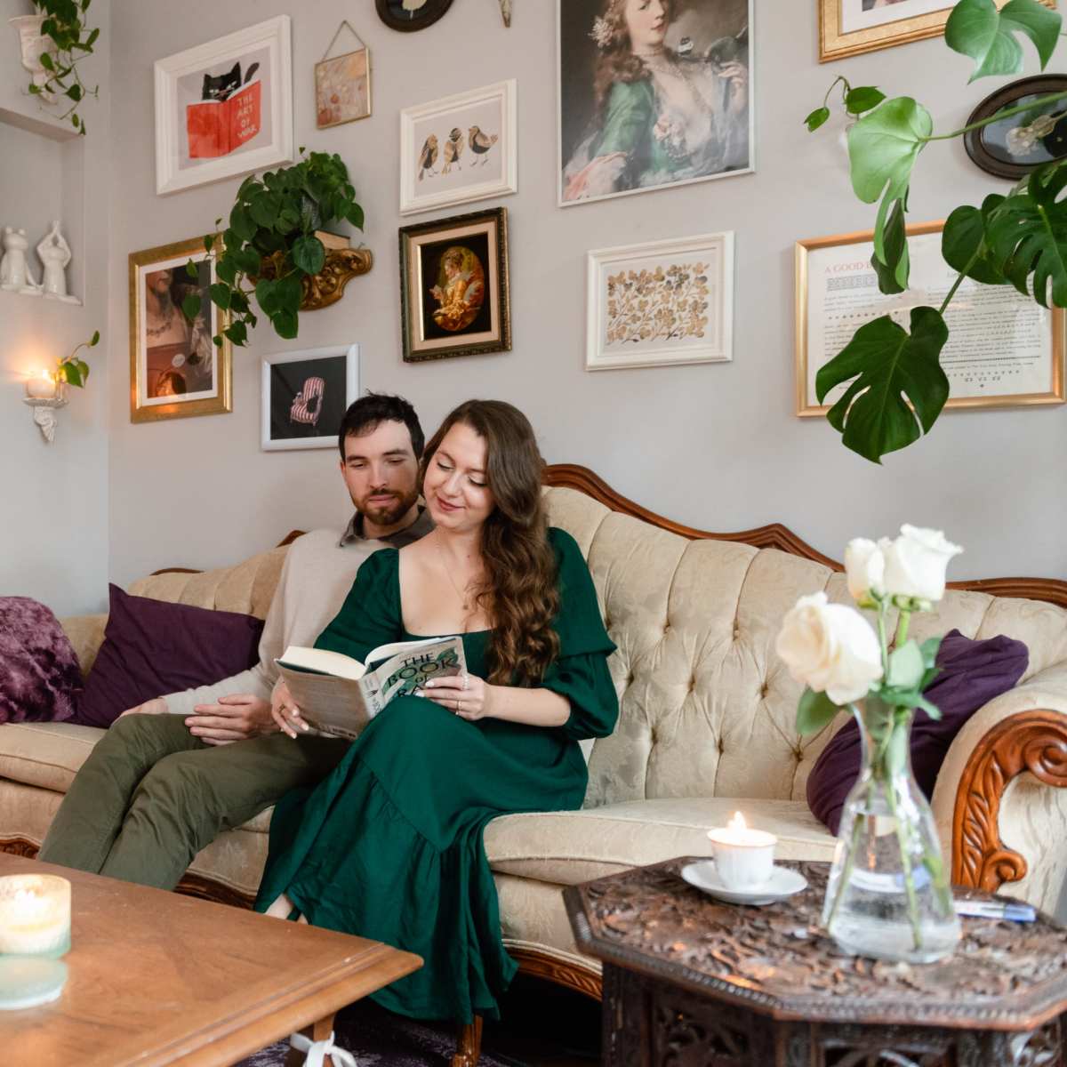 A couple sits together on a vintage couch in a cozy living room, reading a book while surrounded by plants and framed artwork.
