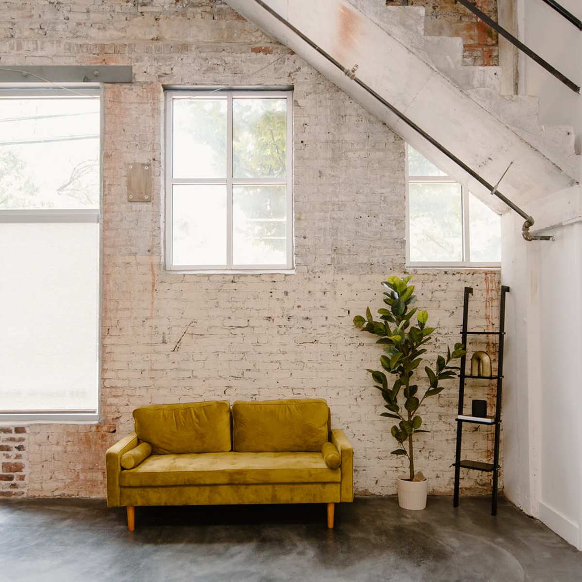 A spacious room with a yellow couch, a potted plant, and large windows, alongside a staircase and exposed brick walls.