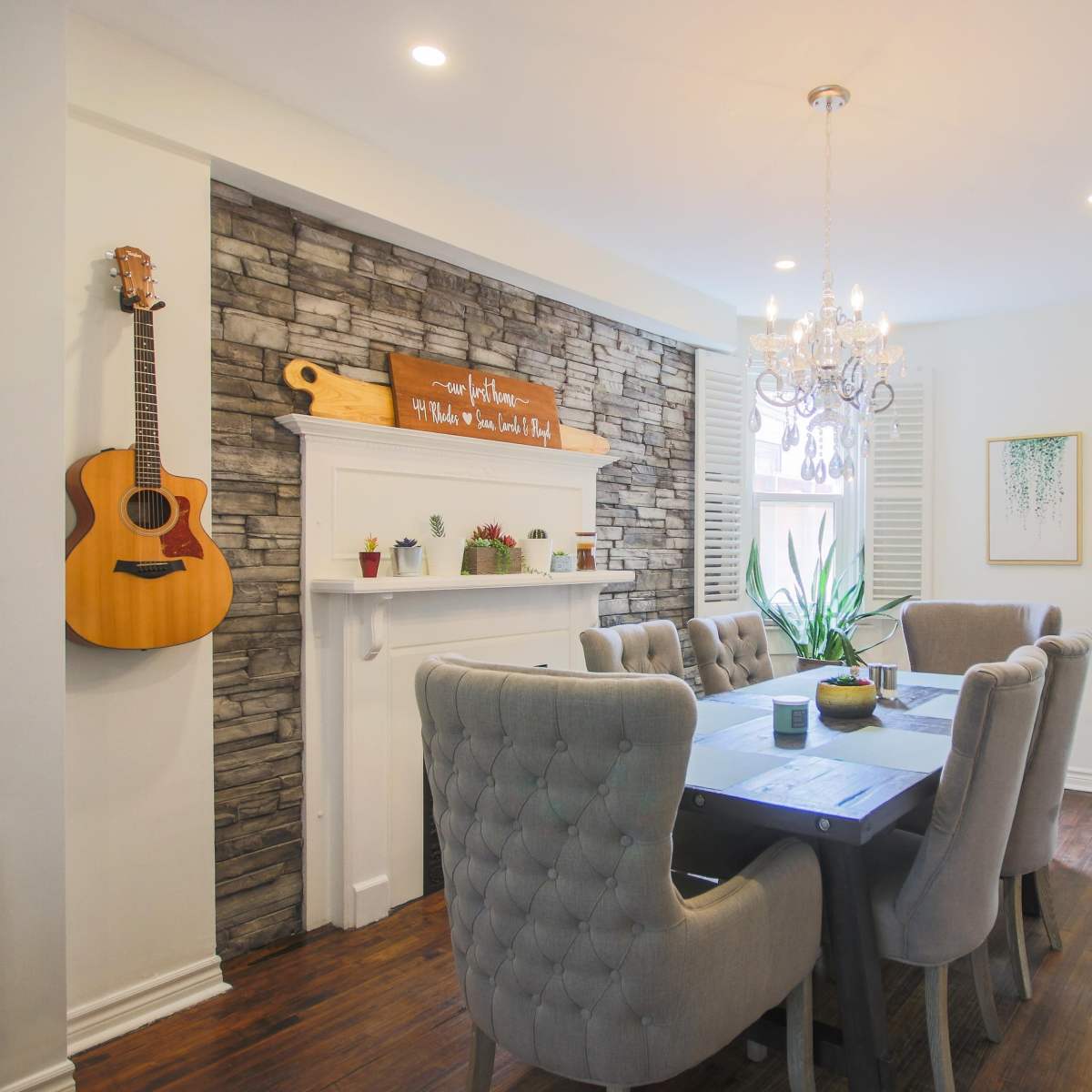 A dining area features a stone accent wall, a large gray table surrounded by upholstered chairs, and a chandelier above, with a guitar hanging on the wall.