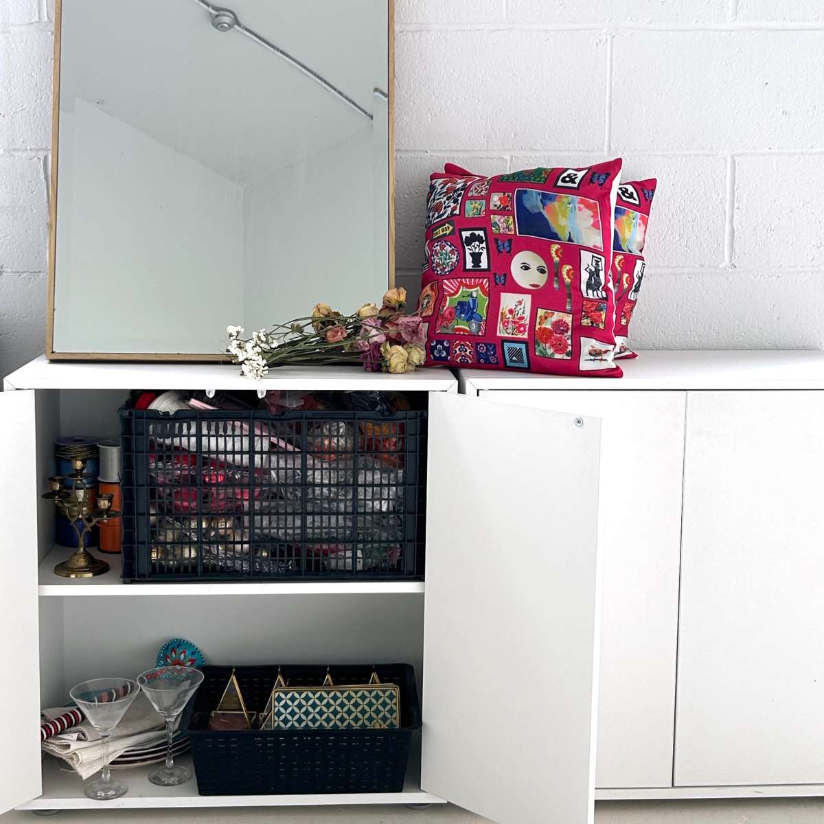A white cabinet with open doors displays various items, including a black basket and a collection of glassware, while a mirror and a decorative bag sit above it.