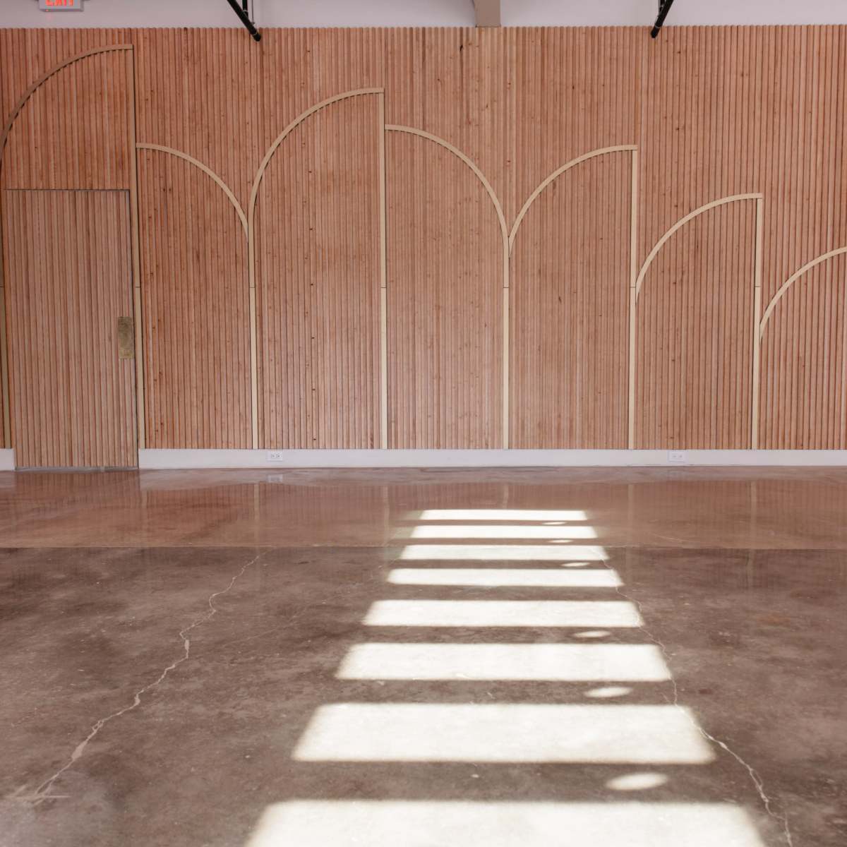 A spacious interior with a wooden paneled wall featuring an arched design and concrete flooring reflecting strong sunlight.