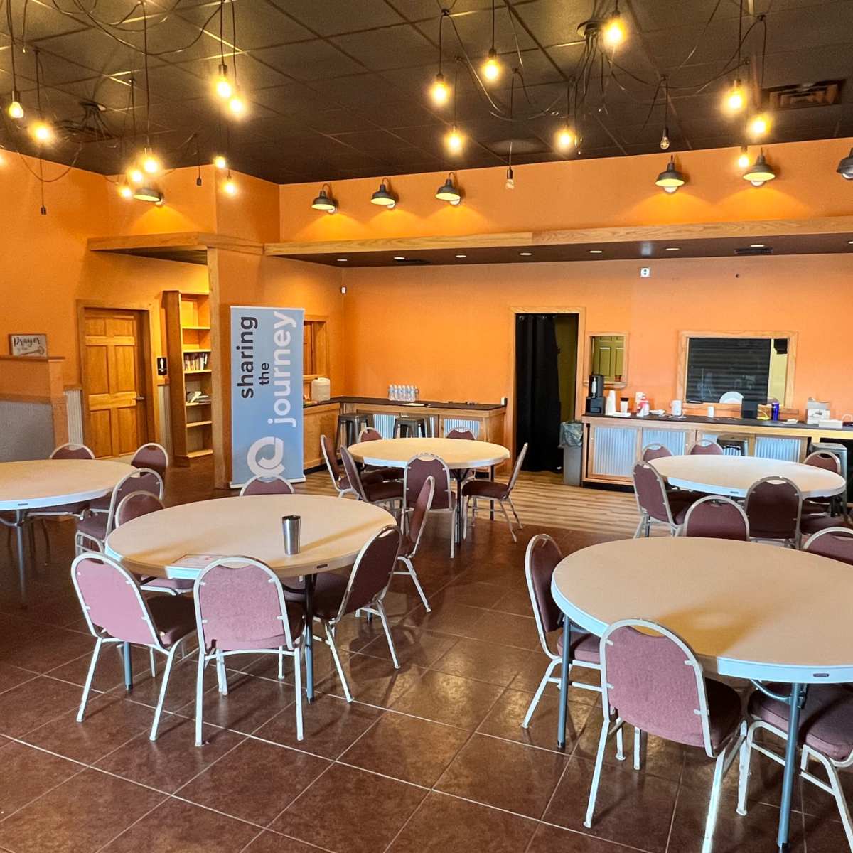 A restaurant-style interior with several round tables surrounded by chairs, an orange-painted wall, and a service counter in the background.