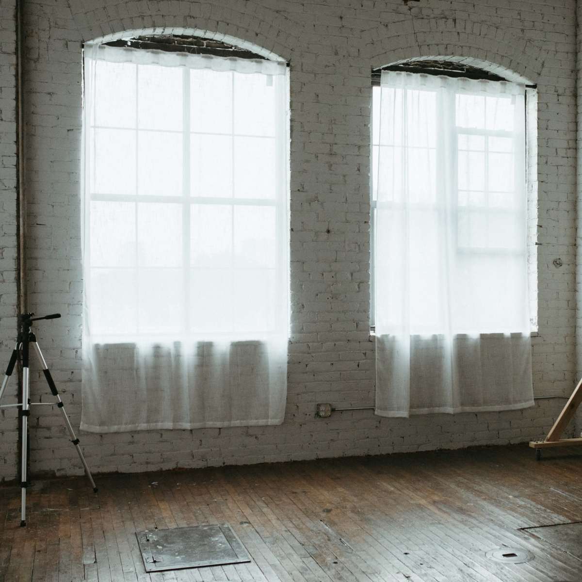 A minimalistic room with large windows draped in sheer white curtains, a large dark panel leaning against the wall, and a tripod standing nearby on the wooden floor.