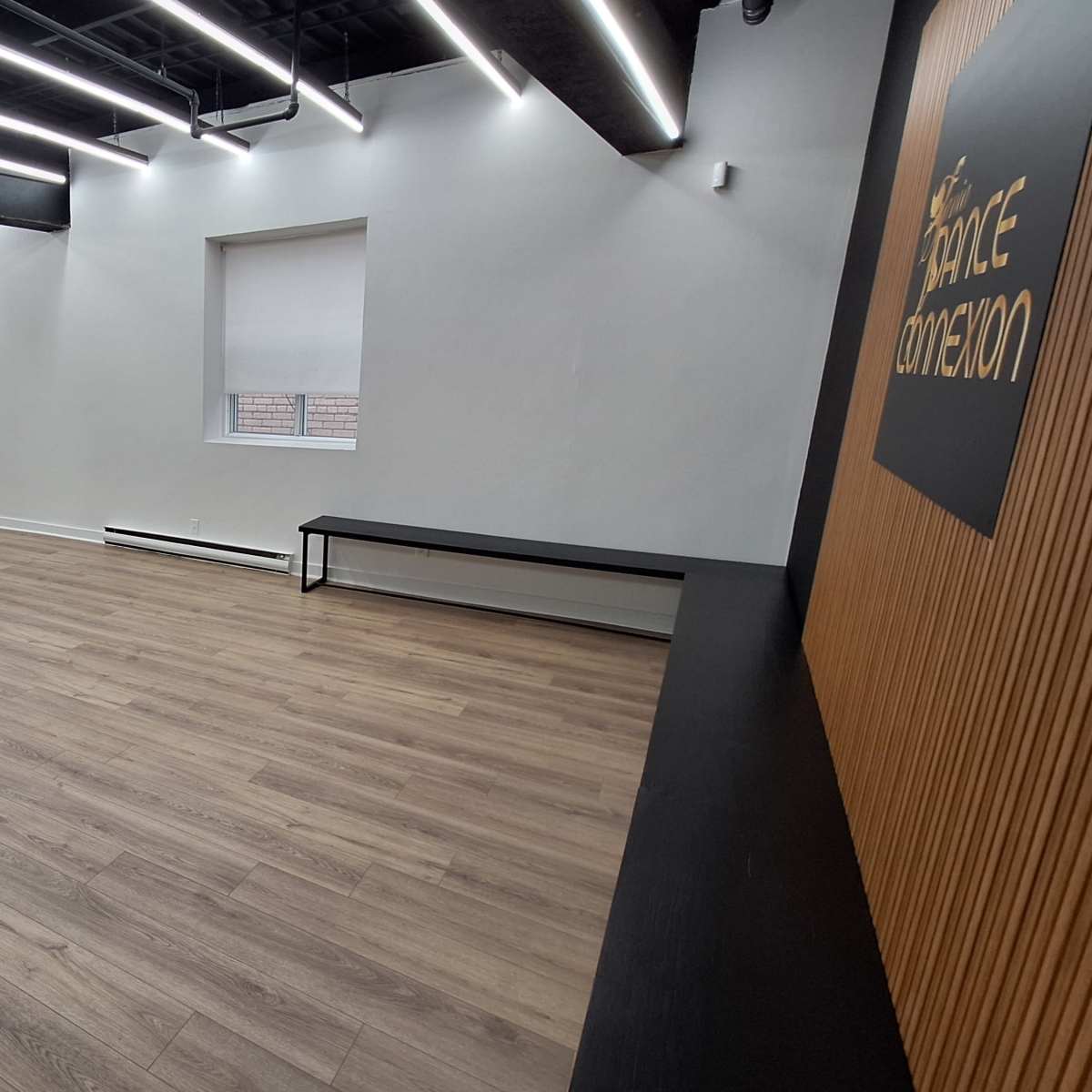 A spacious, empty room with wooden flooring, long ceiling lights, and a wooden wall with a logo on it.
