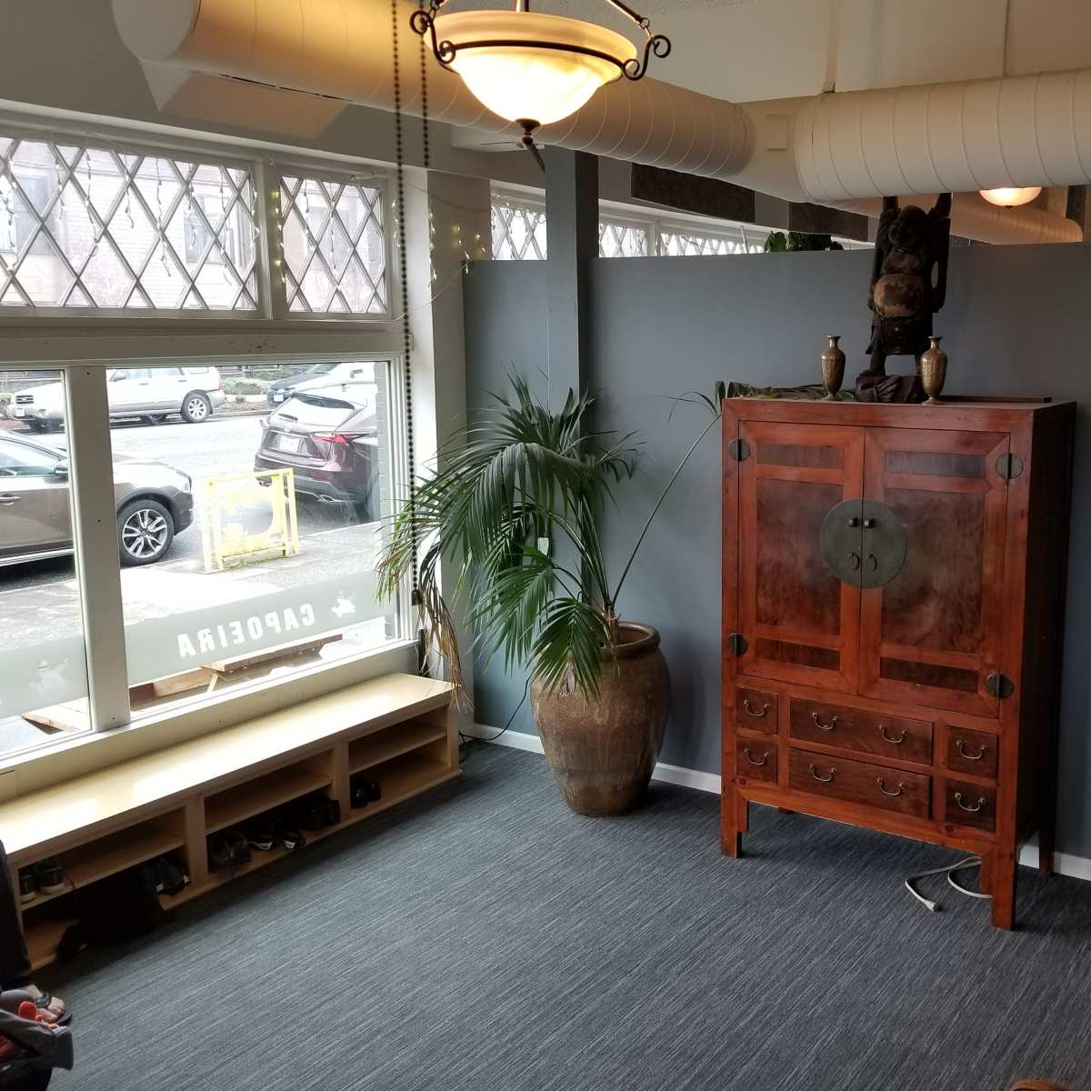 A spacious interior with a wooden cabinet against a gray wall, flanked by potted plants and a shoe rack, featuring large windows that overlook a street.