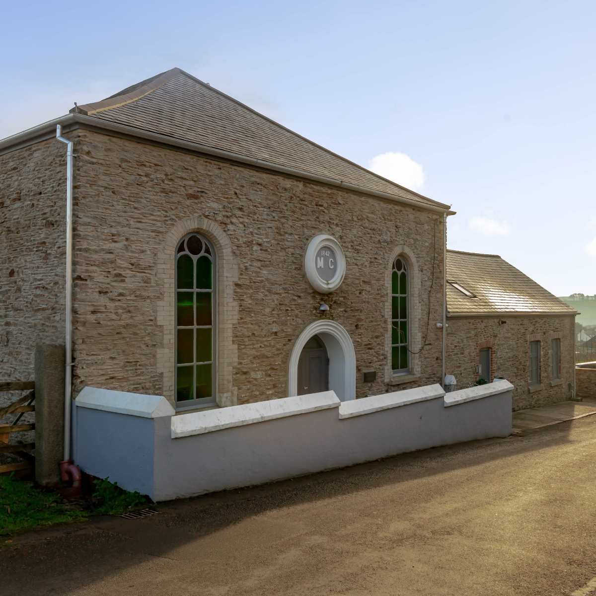 A stone building with arched windows and a sloped roof, situated beside a gravel road surrounded by greenery.
