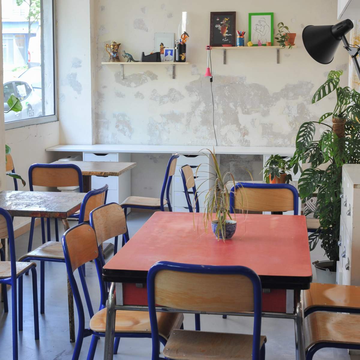 A light-filled cafe interior with wooden tables and chairs, a large window, and shelves displaying decorations.