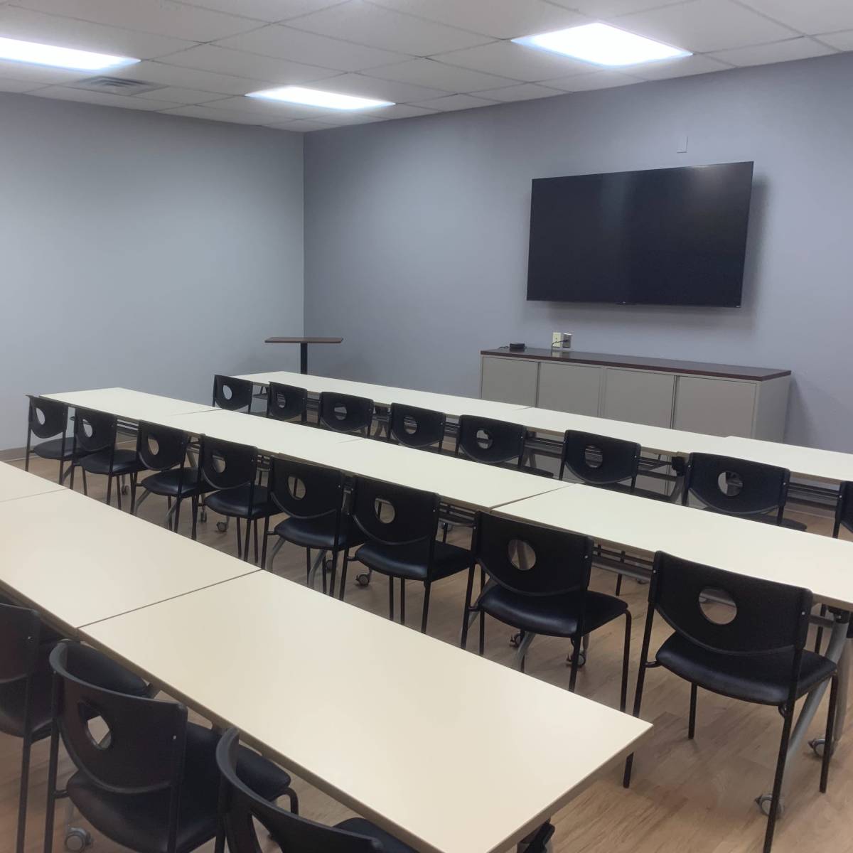 A classroom or meeting room with several rows of tables and black chairs facing a wall-mounted television.