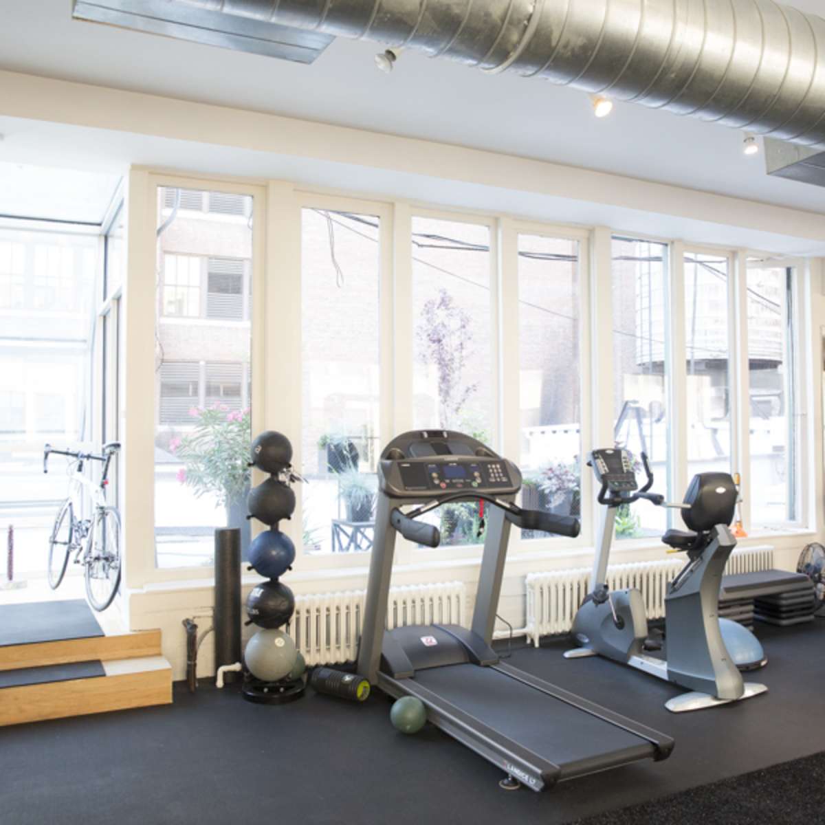 A fitness room features a treadmill, stationary bike, exercise balls, and free weights, with large windows allowing natural light to enter.