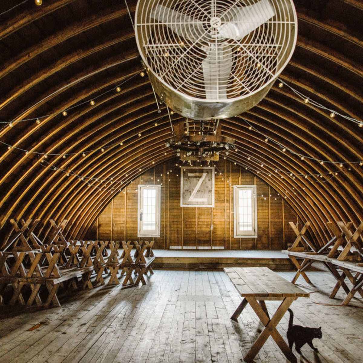 The interior of a rustic barn features arched wooden beams, a large ceiling fan, and a series of tables arranged in rows along the floor.