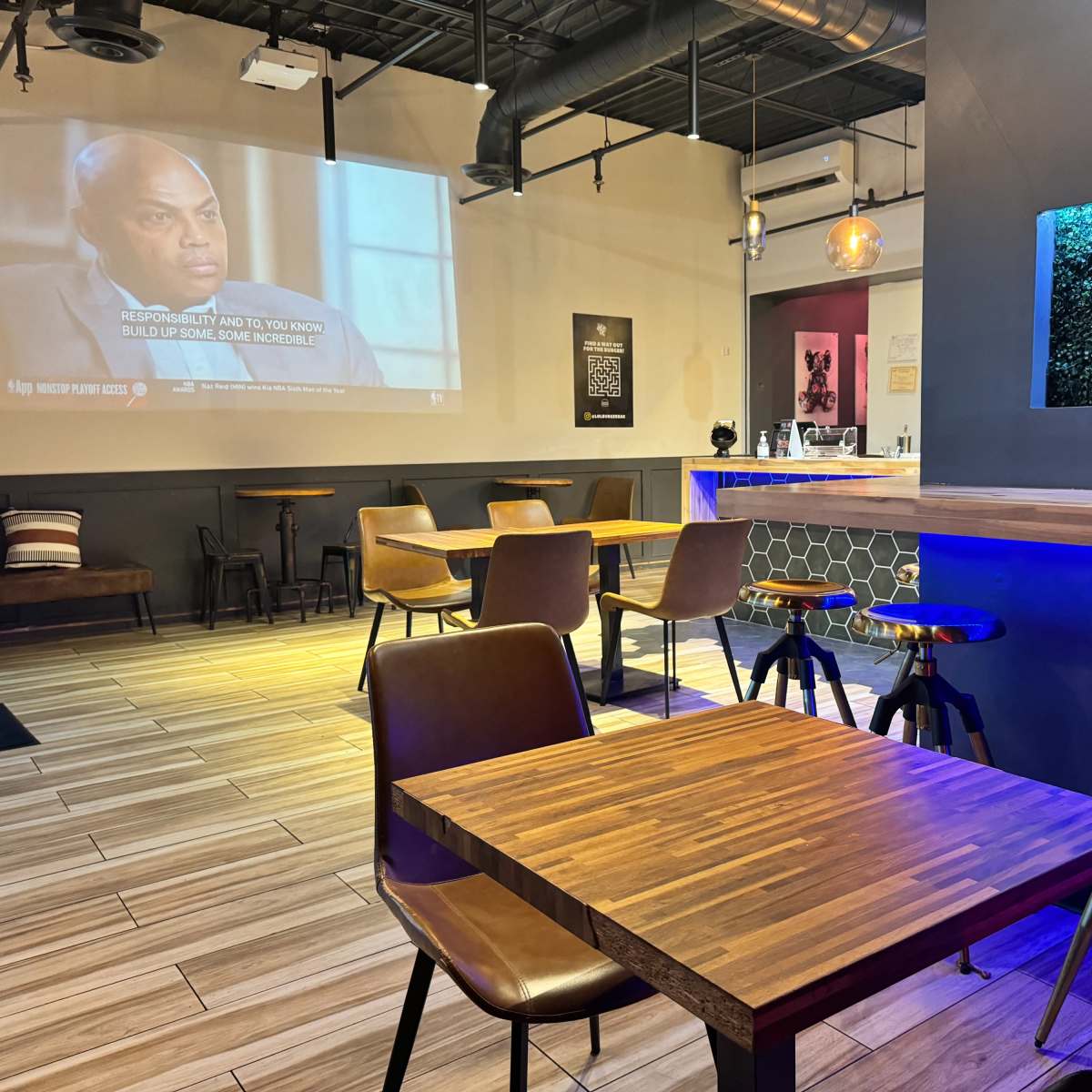 An empty café interior with wooden tables and chairs, a wall-mounted screen displaying a program, and a blue-lit counter area.