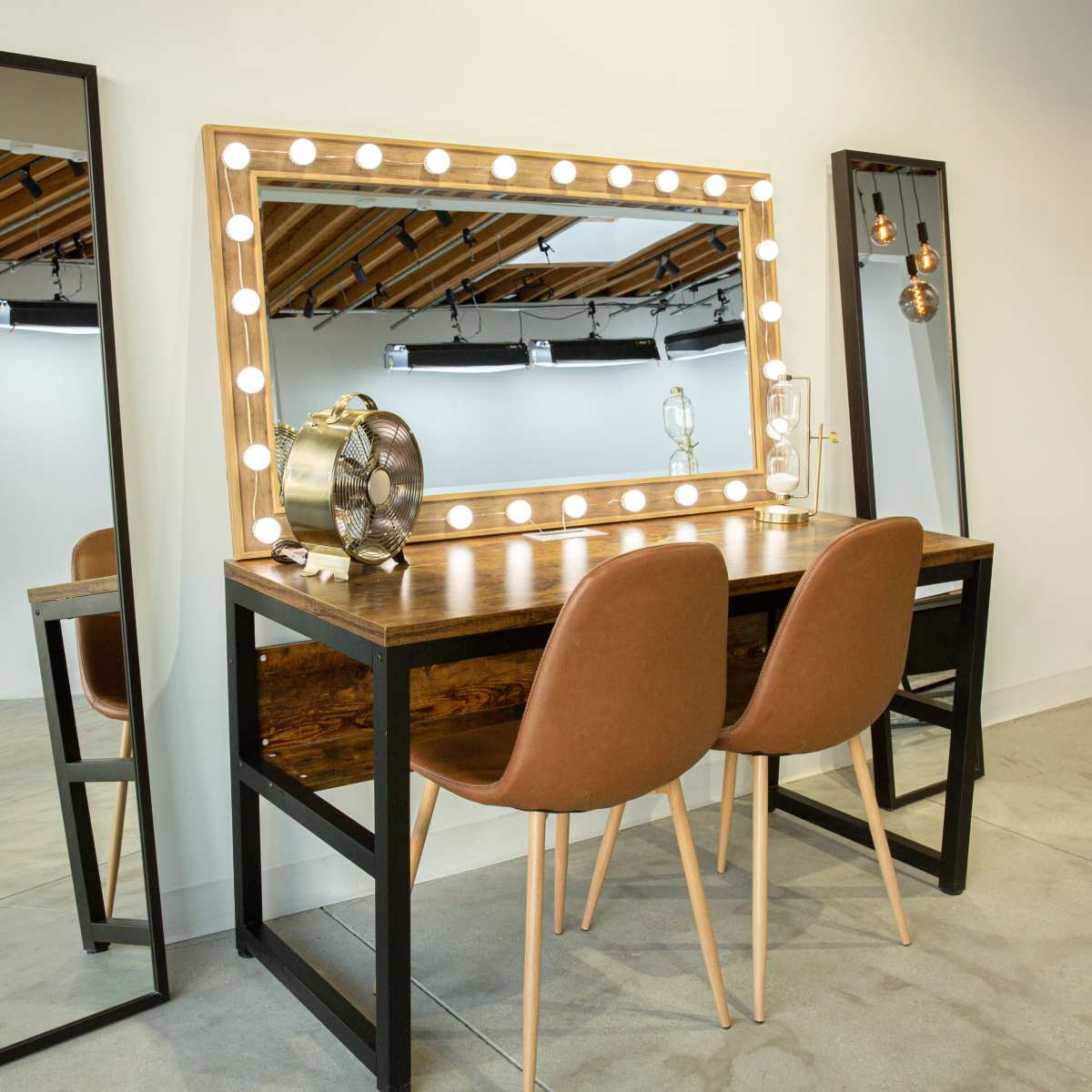 A well-lit vanity area featuring a wooden table with two chairs, surrounded by large mirrors framed with lights.