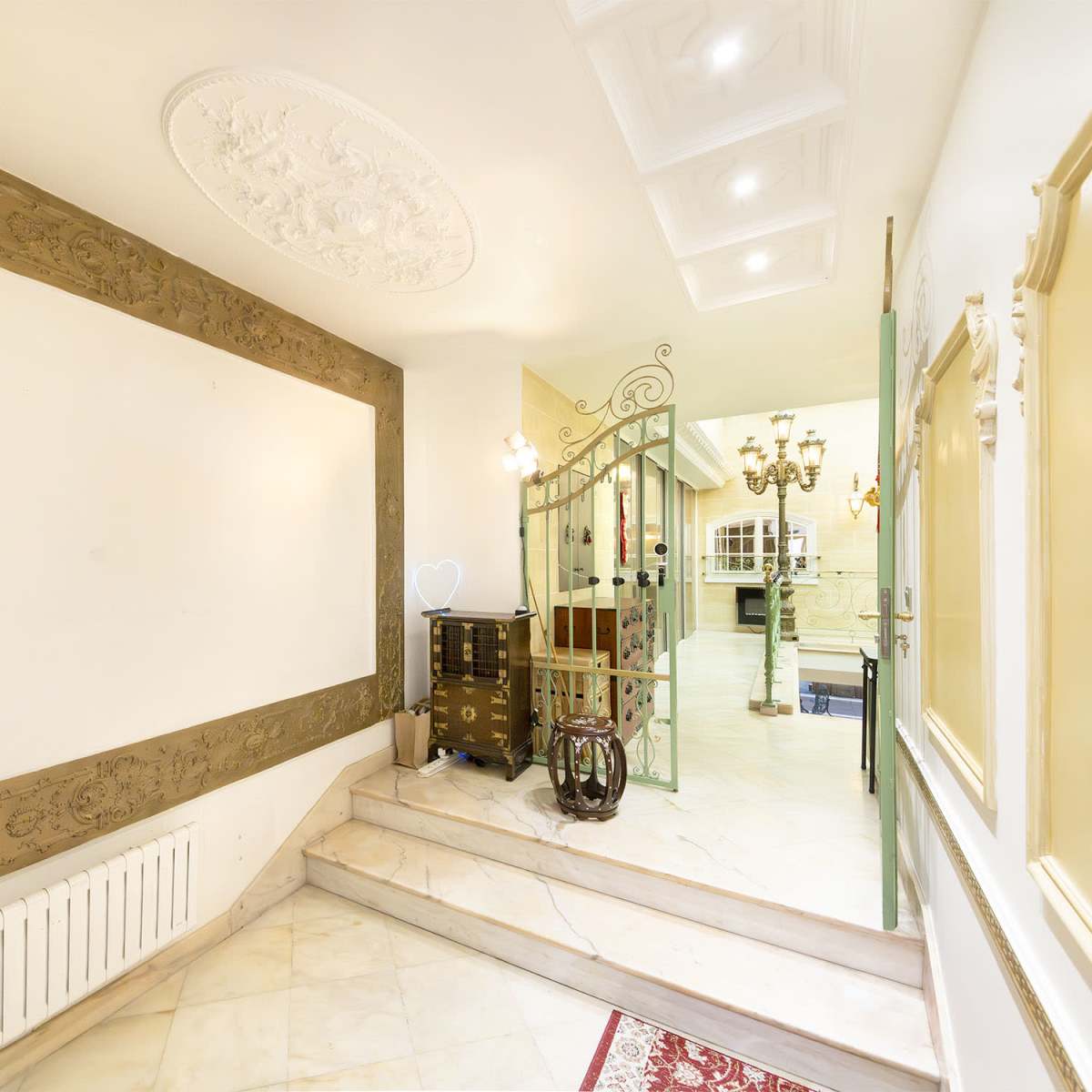 A well-lit hallway featuring decorative molding on the walls, a staircase leading to another area, and an ornate gate leading to a room with chandeliers.