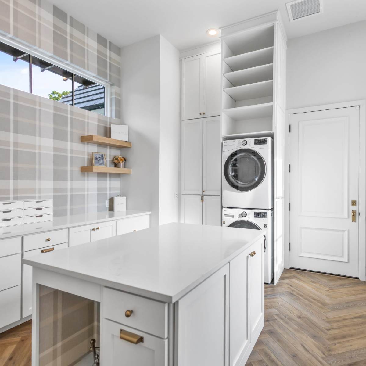 A modern laundry room with white cabinetry, a stacked washer and dryer, and a large central island on a herringbone-patterned floor.