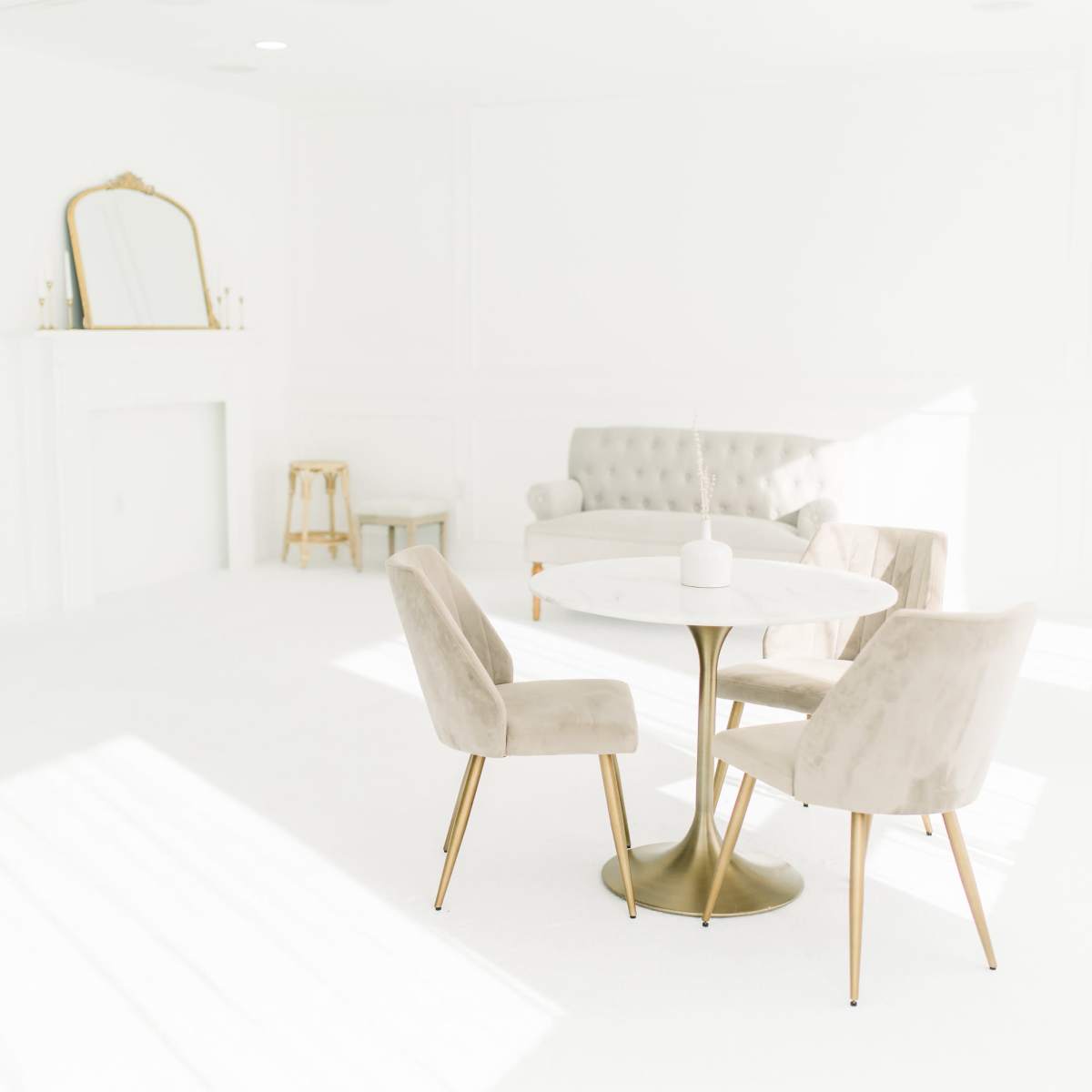 A minimalist living space features a round table surrounded by four beige chairs, a light-colored couch, and a fireplace with a large mirror above it.