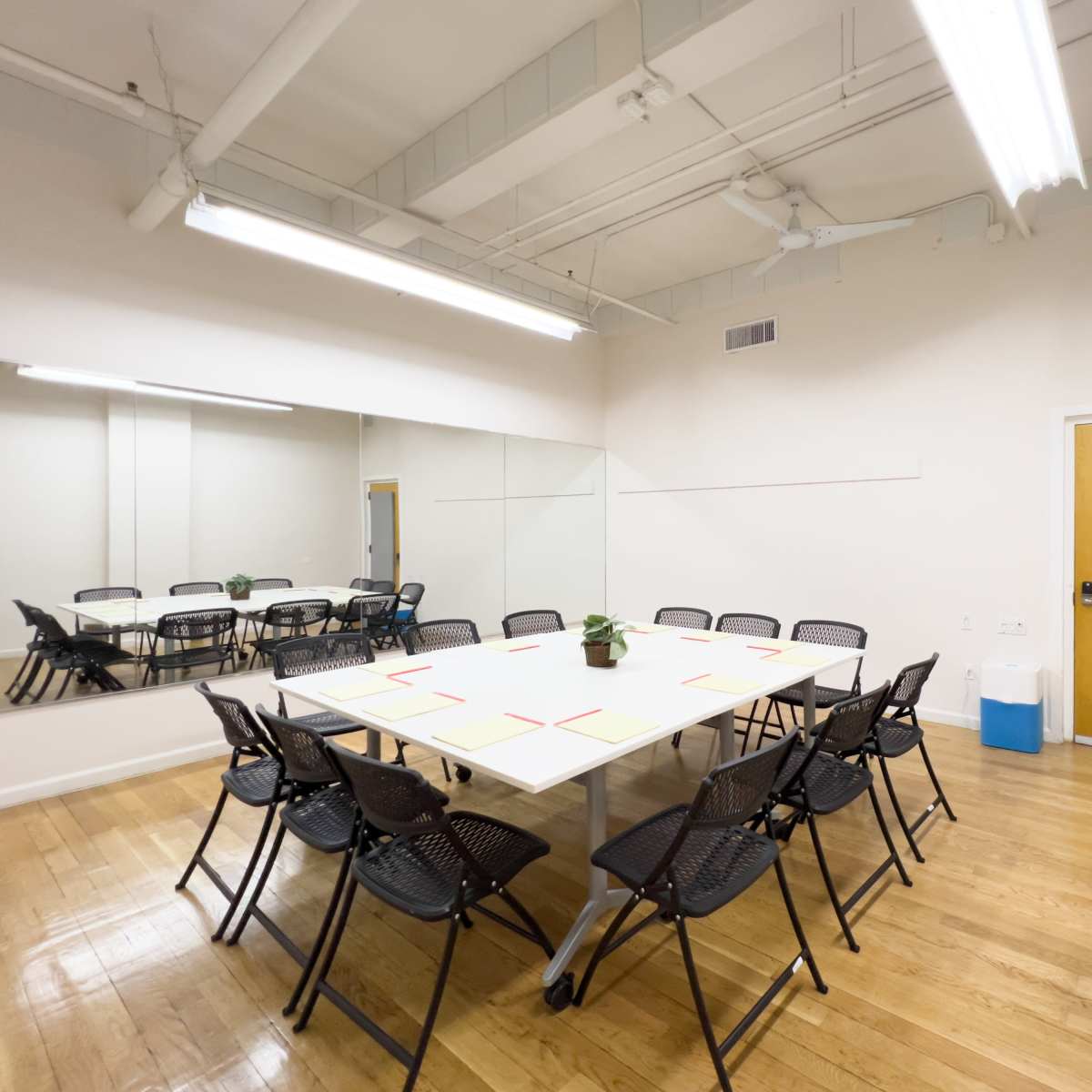 A meeting room features a rectangular table surrounded by black chairs, with a mirror on one wall and a door on another.