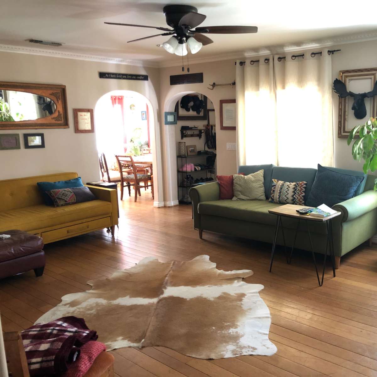 The living room features a yellow sofa, a green couch, a coffee table, a cowhide rug, and large windows with light curtains.
