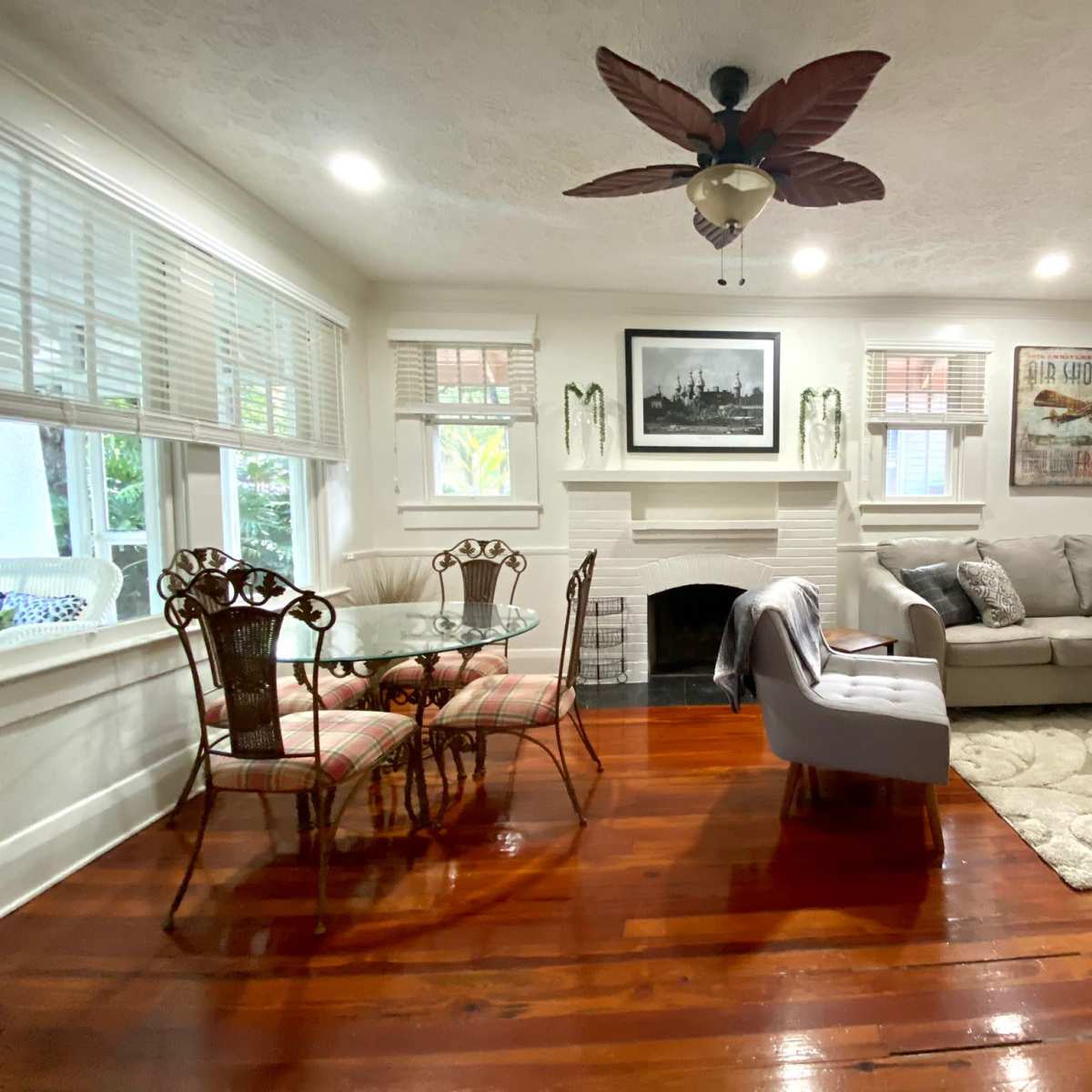 A living room with a sofa, a round dining table, a fireplace, and large windows, all set against hardwood flooring.