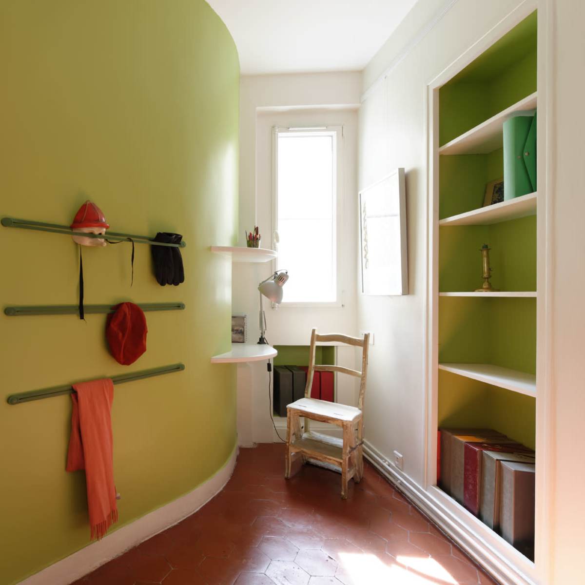 A small room with a bright green curved wall, a sink, shelves on one side, and a wooden chair near a window.