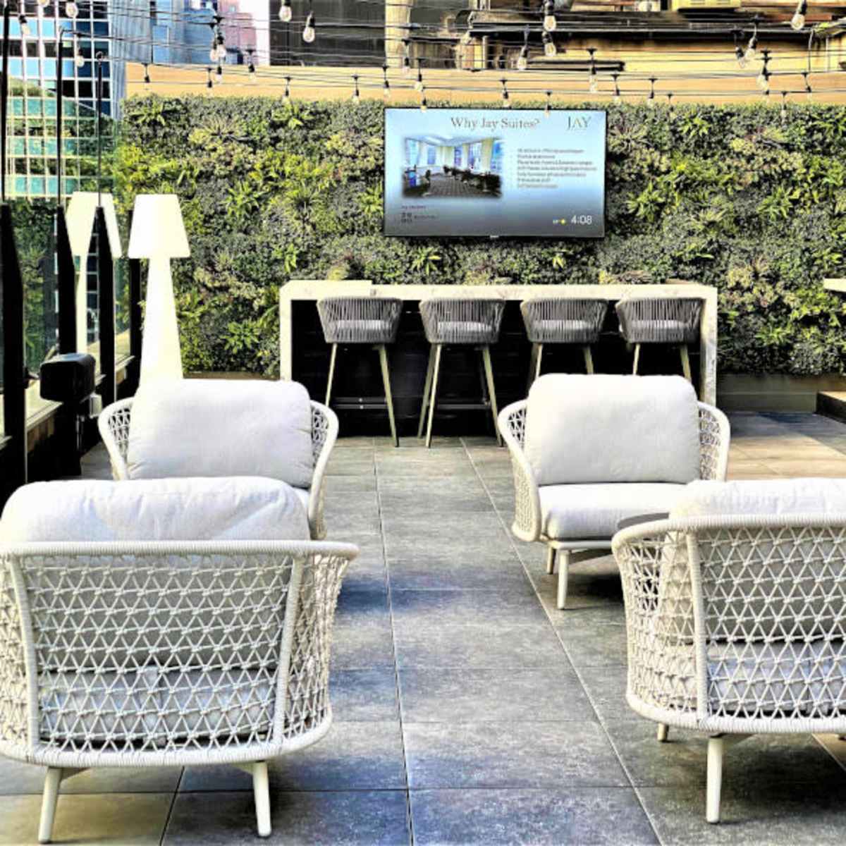 A modern rooftop lounge area with four white wicker chairs arranged around a small table, a wall of greenery behind, and a digital display mounted on the wall.