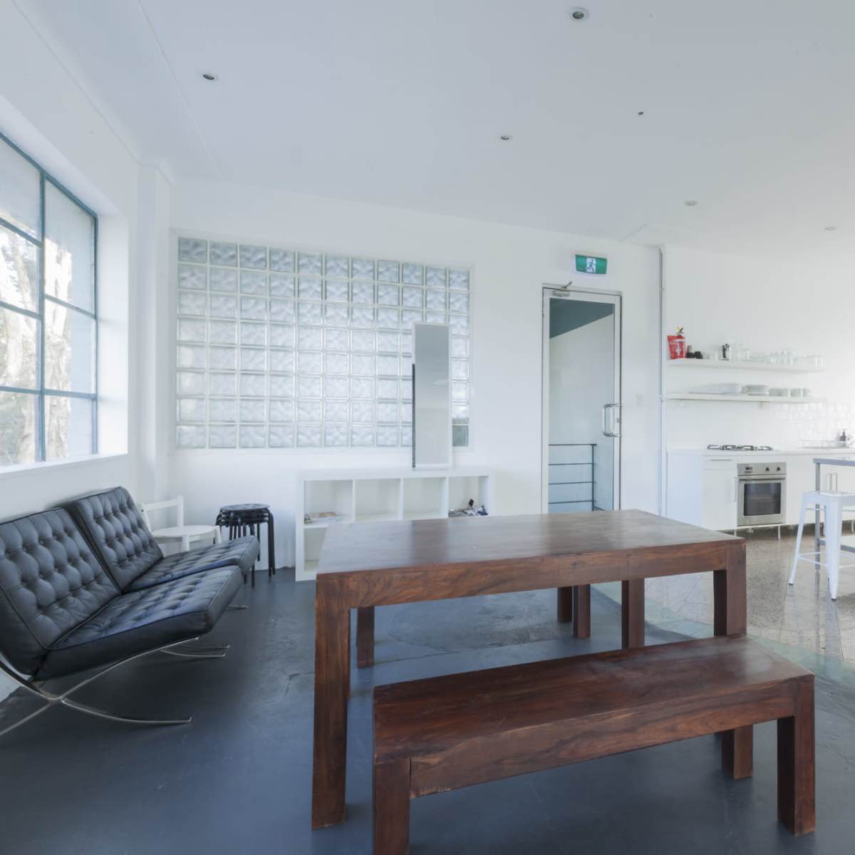 A modern, minimalist kitchen and dining area with a wooden table, a black sofa, and large windows allowing natural light.