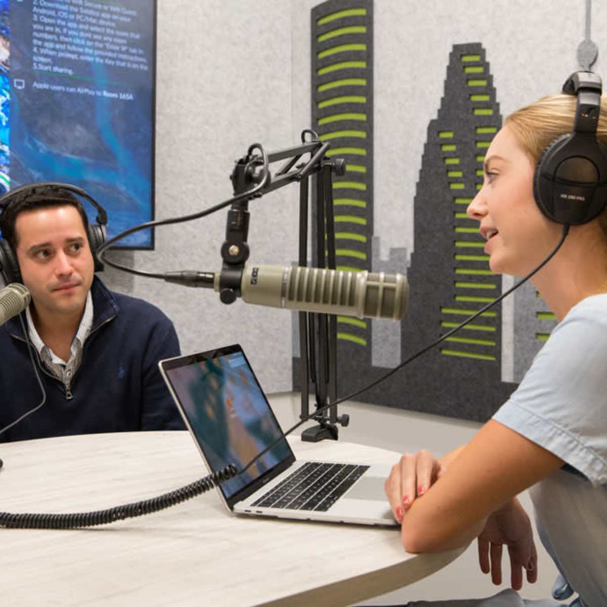 A man and a woman are engaged in a conversation at a table in a recording studio, equipped with microphones and a laptop.