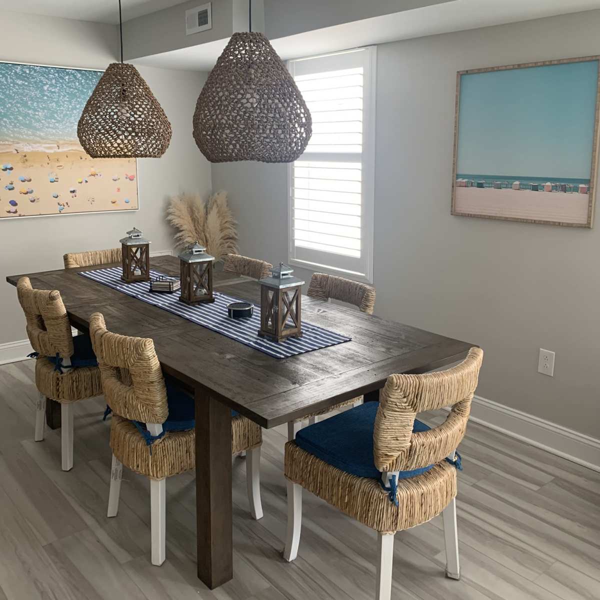 A modern dining room features a wooden table surrounded by woven chairs, two large pendant lights, and artwork on the walls.