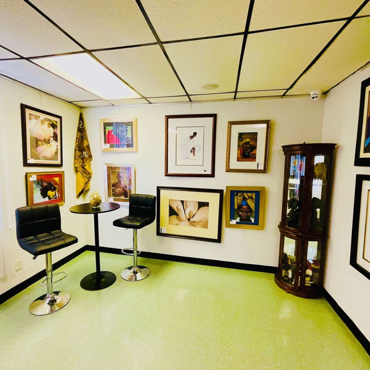 A small gallery room features a set of framed artworks on the walls, a round table with two black stools, and a display cabinet in one corner.