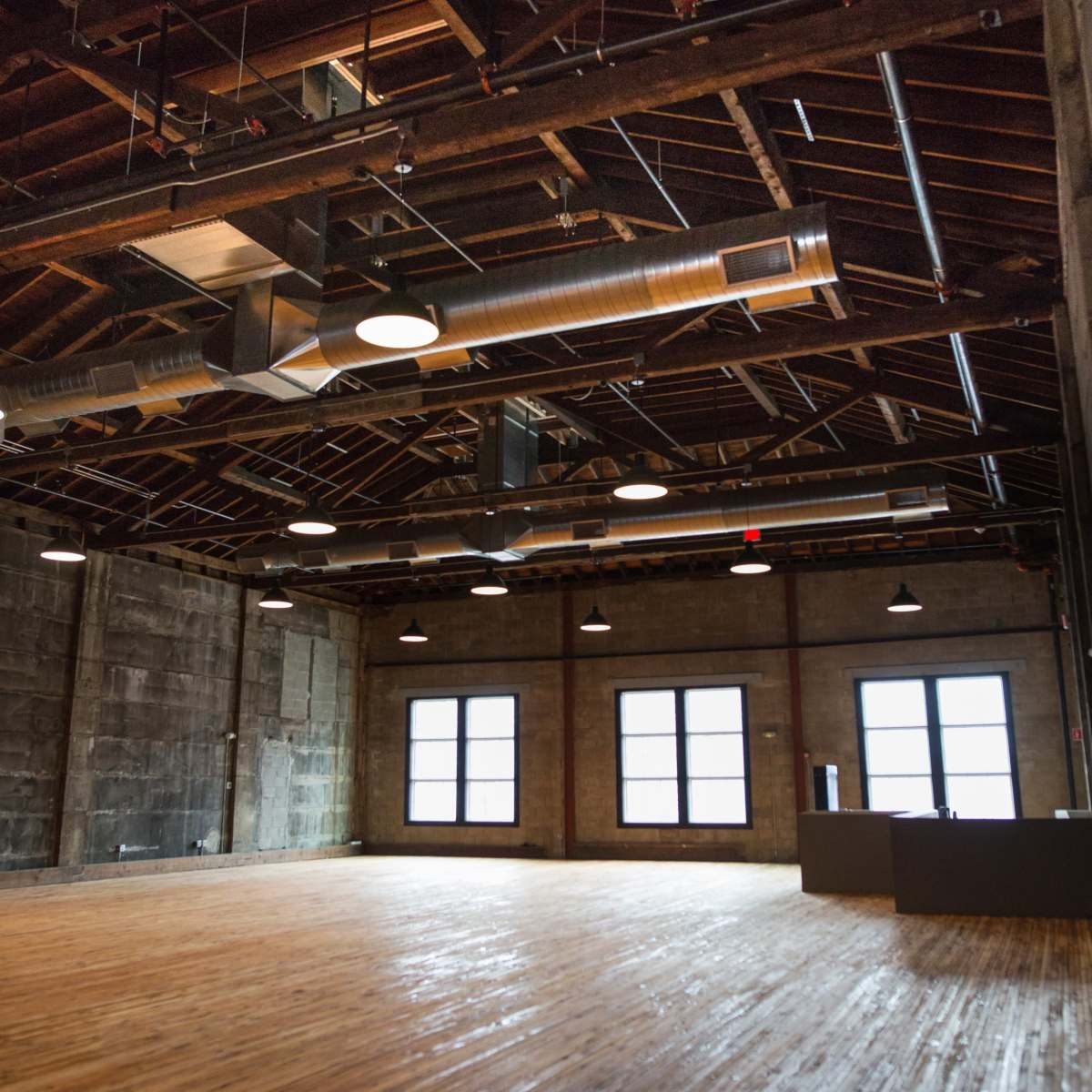 An empty industrial-style room with wooden floors, exposed beams, and large windows.