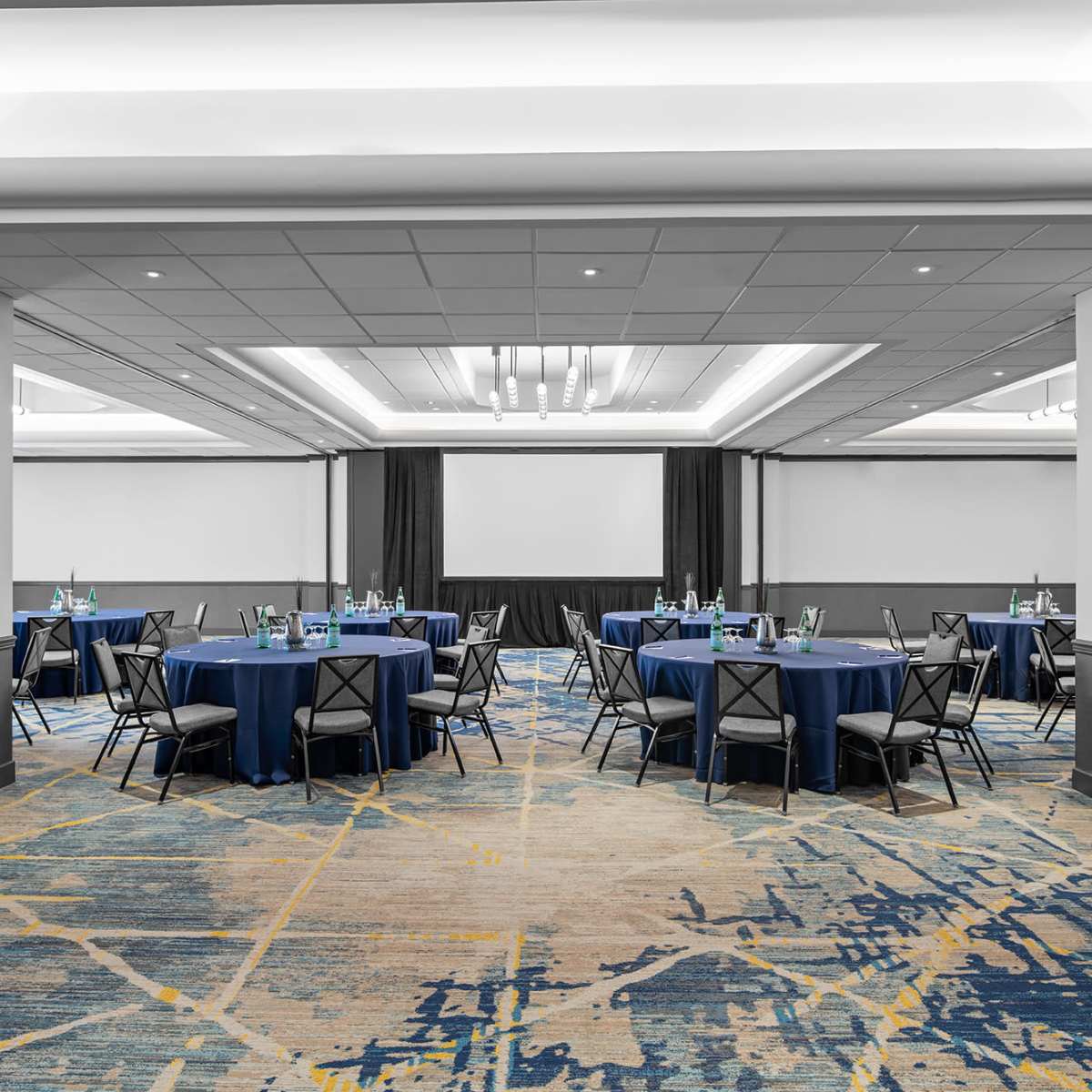 A spacious event room features multiple round tables dressed in blue tablecloths, surrounded by black chairs, with a large projection screen at the front.
