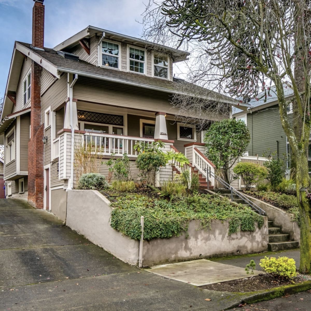 A large, two-story home with a front porch is situated on a sloped driveway, surrounded by landscaped greenery and trees.