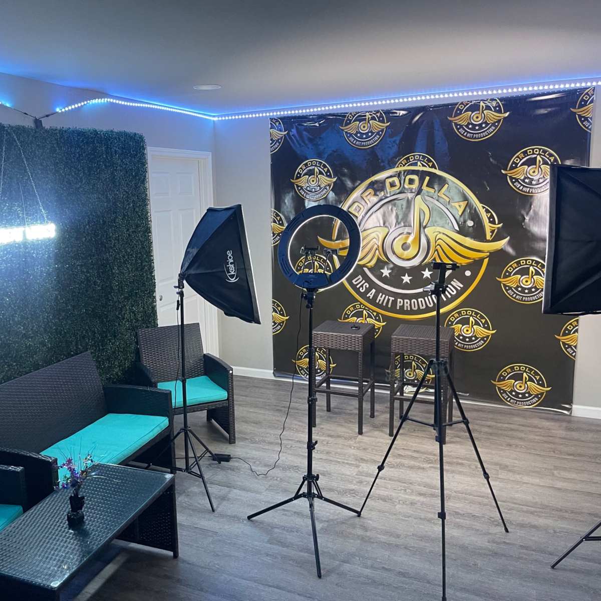A modern recording studio setup with green walls, a backdrop featuring a logo, and lighting equipment arranged around seating.