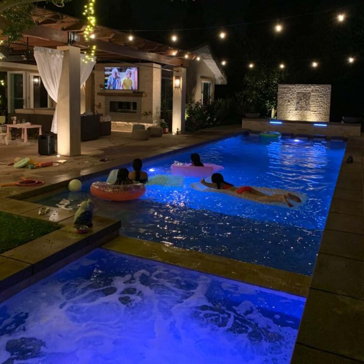 The scene depicts a backyard pool area at night, illuminated by string lights and featuring people swimming while a television displays a show nearby.