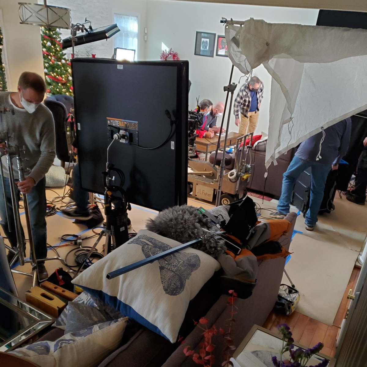 A film crew sets up equipment in a living room decorated for the holidays, featuring a Christmas tree and various filming gear scattered around.