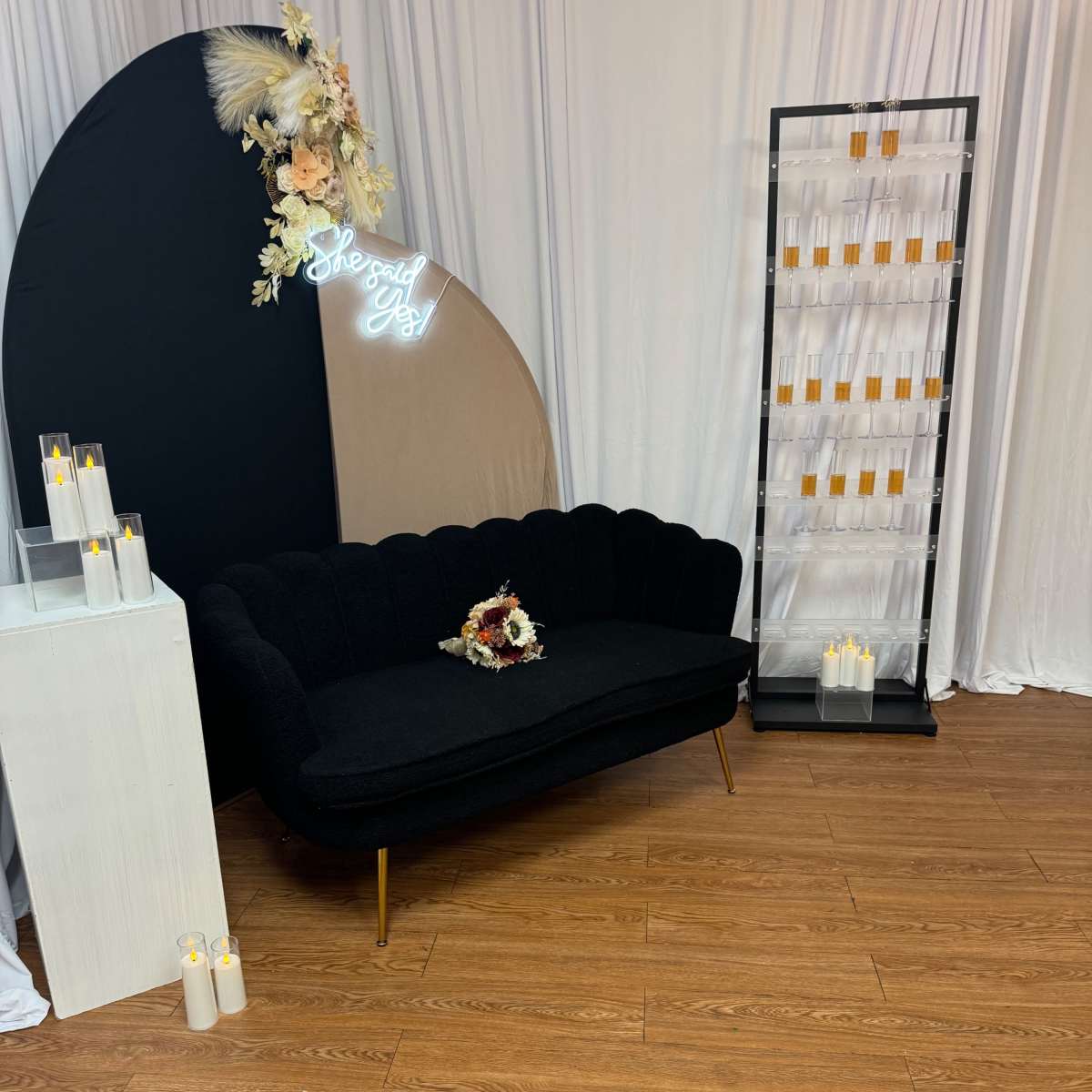 A black tufted couch placed against a backdrop curtain, accompanied by a floral arrangement and a display of candles on a stand.