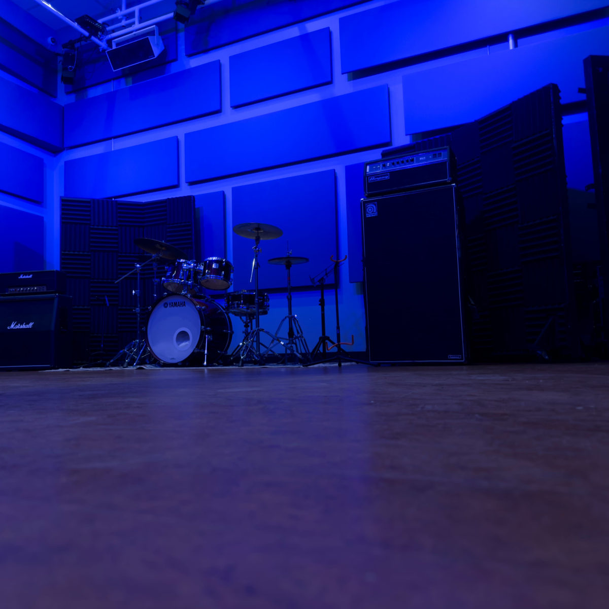 A music practice room featuring a drum set, amplifiers, and soundproofing panels under blue lighting.