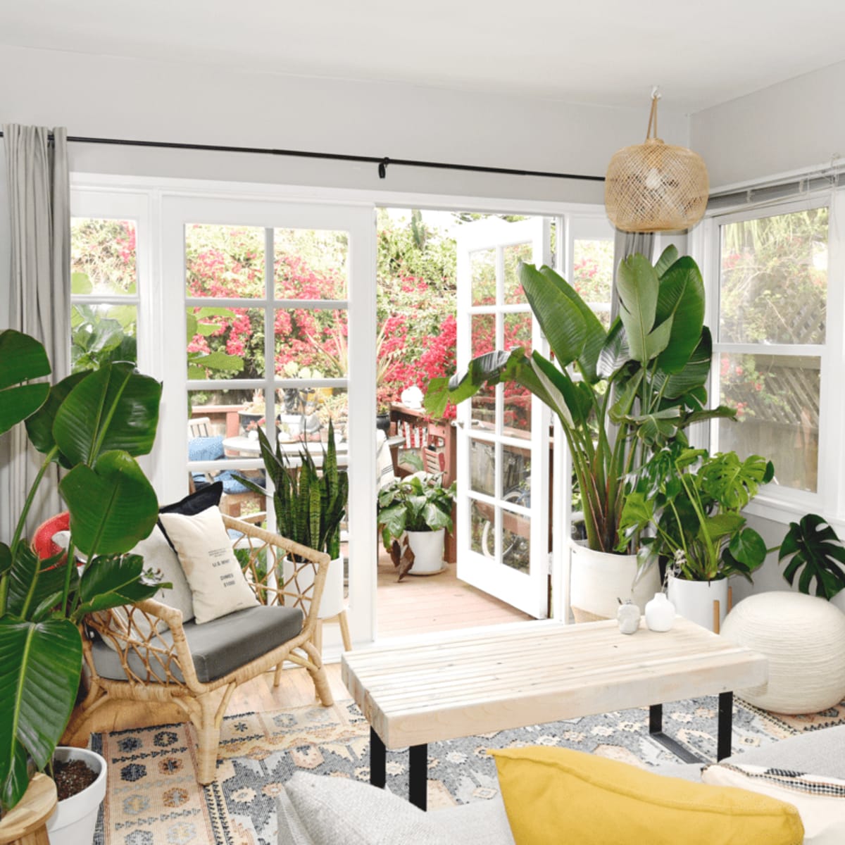 A cozy living room features large windows opening to a garden, surrounded by potted plants and a wooden table in the center.