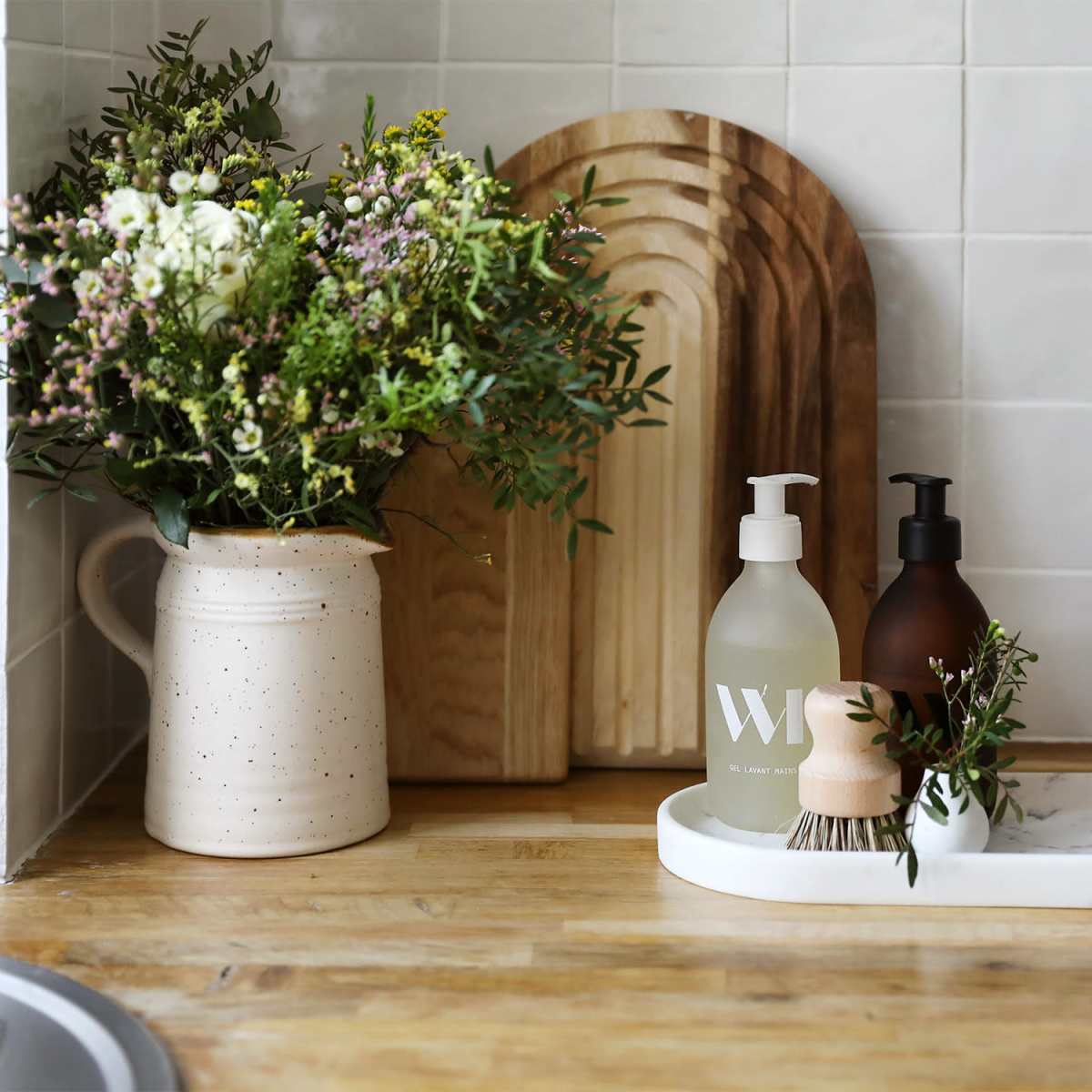 A wooden countertop displays a vase of flowers, two bottles of soap, and a small brush on a marble tray.