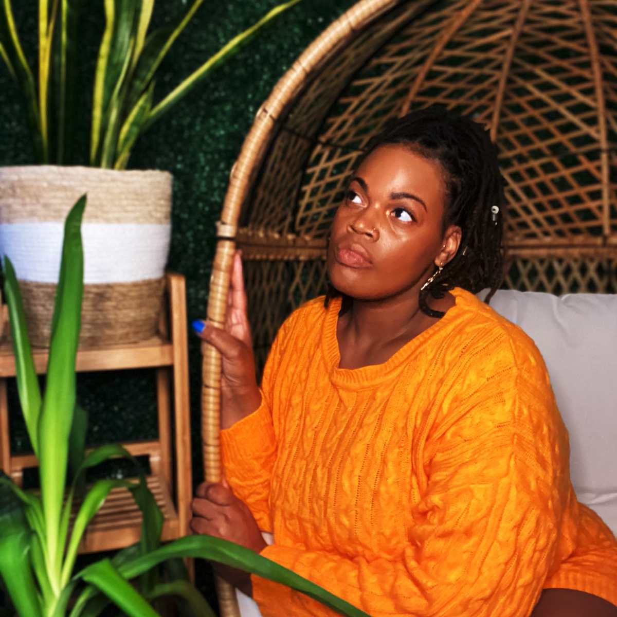 A person wearing an orange sweater sits in a wicker chair surrounded by greenery and plants.