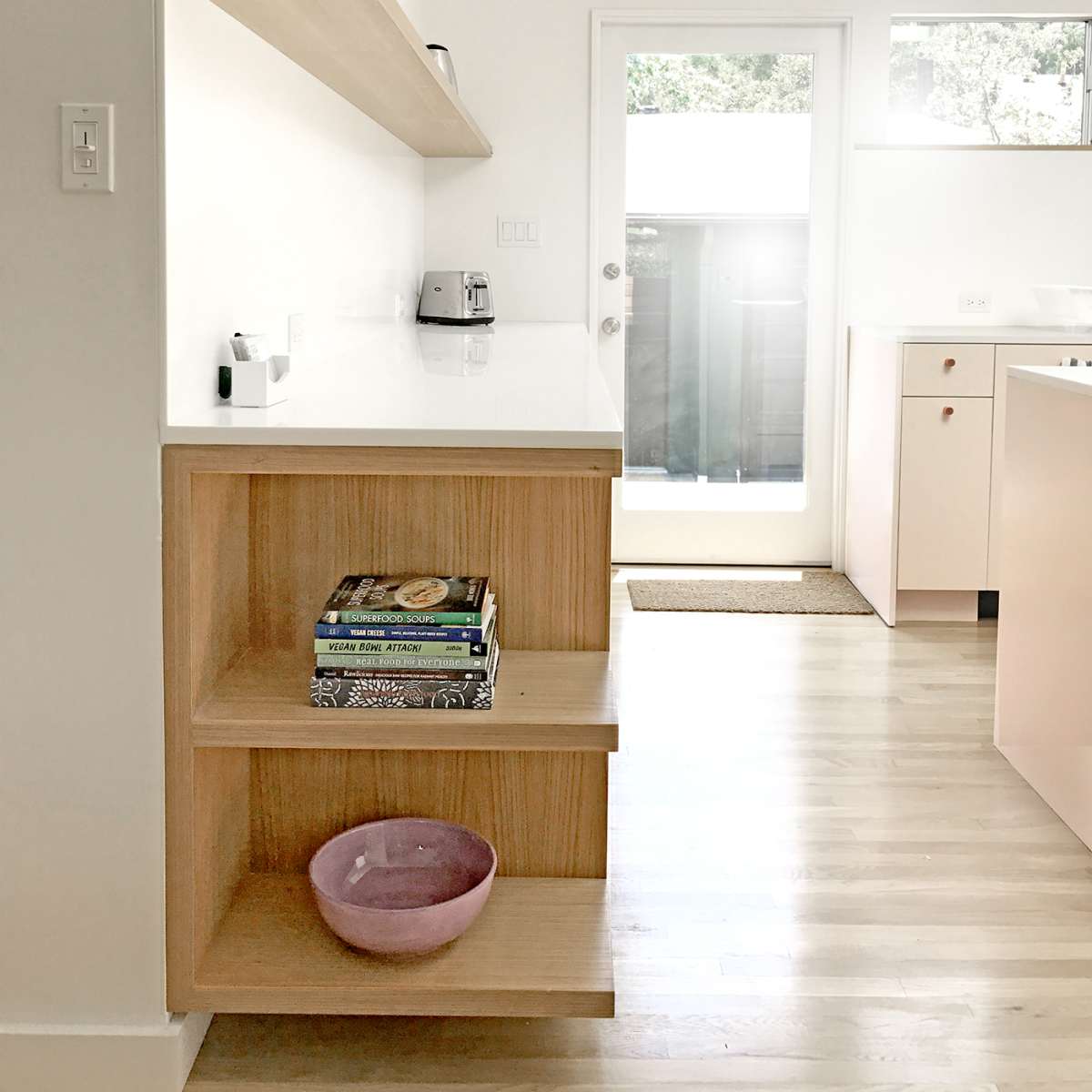 A light-filled kitchen features a wooden shelf with cookbooks and a bowl, adjacent to a sliding glass door leading to an outdoor area.