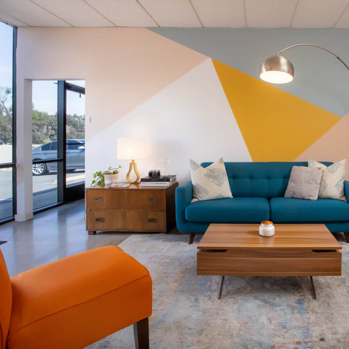 A modern living room with a blue sofa, orange chairs, a wooden coffee table, and a geometric mural on the wall.