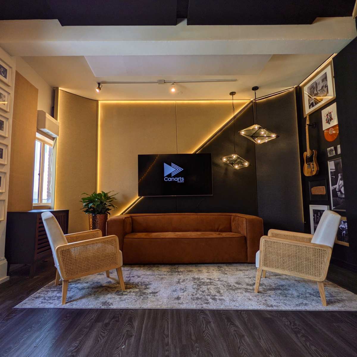 A modern living room with a brown sofa, two light-colored chairs, a television on the wall, and decorative elements including a plant and framed pictures.