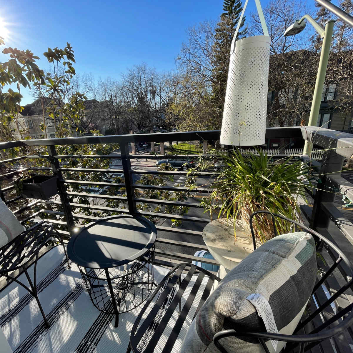 A balcony features two chairs and a small table, surrounded by greenery and overlooking a street.