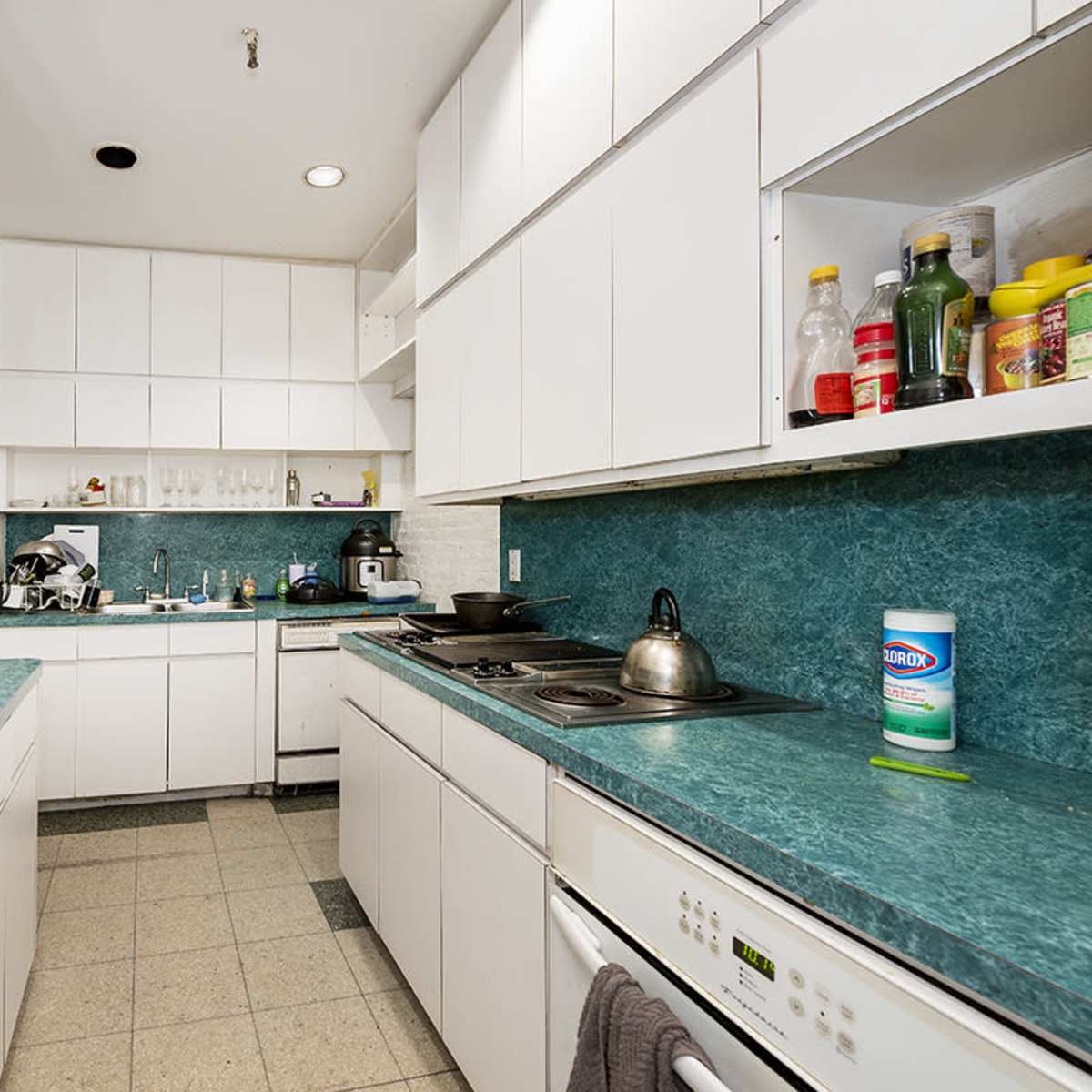 The kitchen features white cabinets, green countertops, and appliances arranged along the walls, with various utensils and condiments on display.