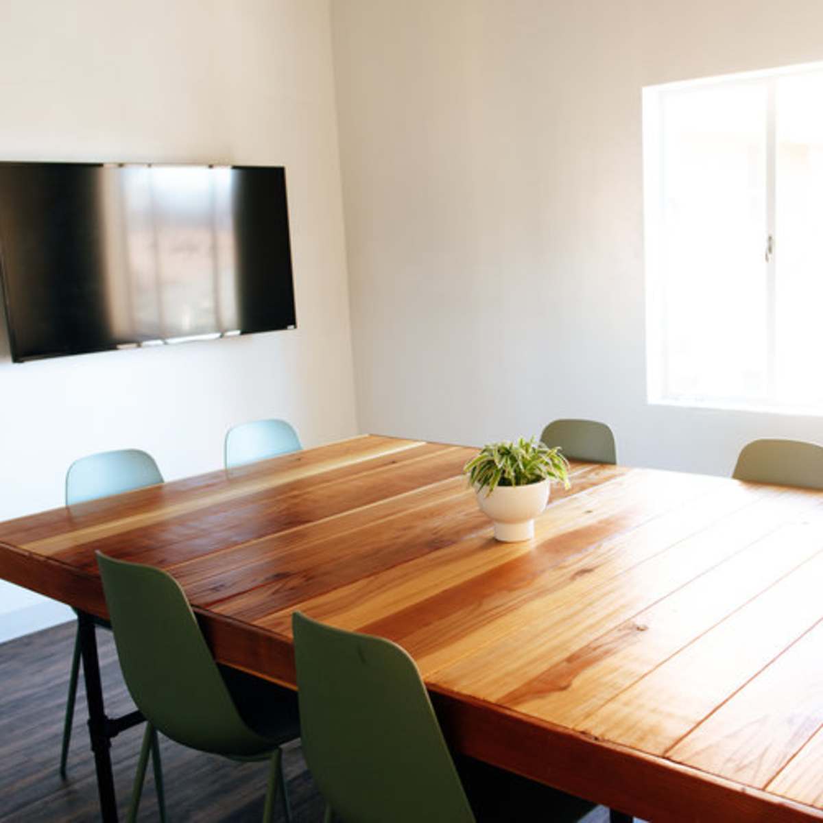 A large wooden table surrounded by chairs is positioned in a brightly lit room with a television mounted on the wall and a window offering an outside view.
