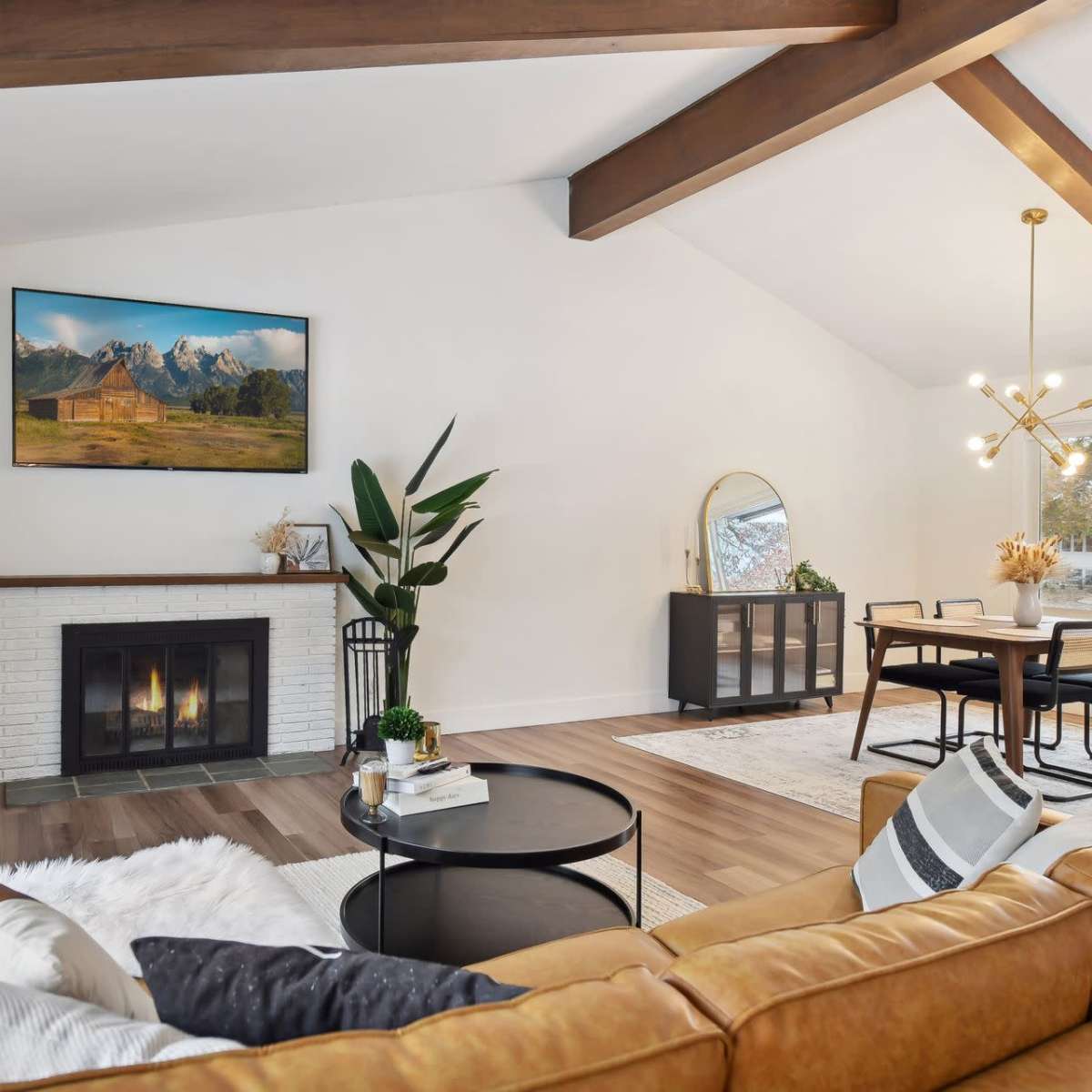 The living room features a cozy seating area with a leather sofa, a coffee table, and a fireplace beneath a large mountain landscape painting, accompanied by a dining space with a round table and modern light fixture.