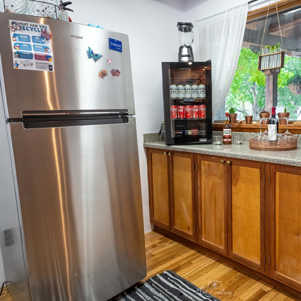 A kitchen corner with a stainless steel refrigerator, a countertop with a variety of drinks in a glass cabinet, and a window with sheer curtains.