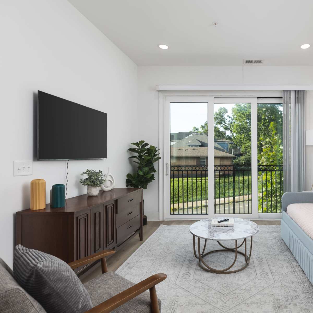 A modern living room features a blue sofa, a wooden console with a television, and a coffee table in front of a large window with a view of greenery outside.