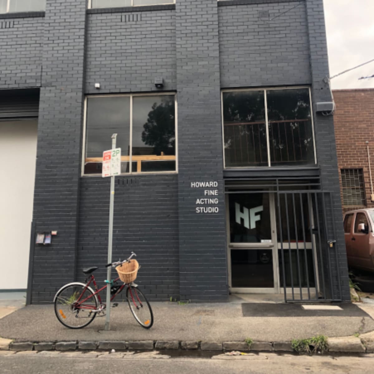A gray brick building with large windows displays the sign "HOWARD FINE ACTING STUDIO," while a bicycle with a basket is parked outside.