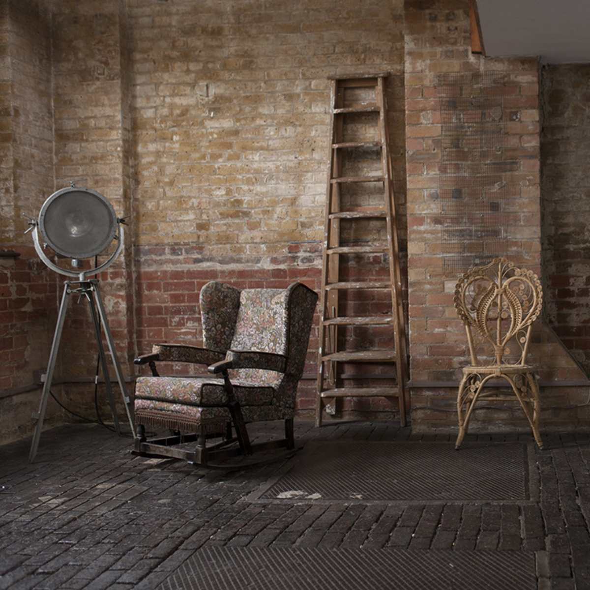 A minimalist interior space with two armchairs, a wooden ladder, a large light fixture, and a mirror against a brick wall.